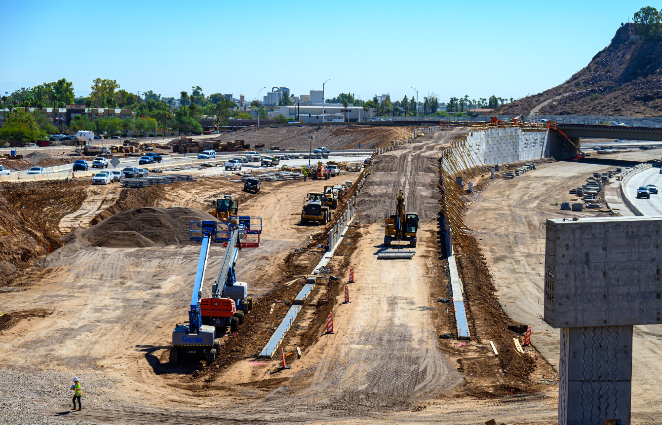 Multiple construction vehicles in the foreground with Interstate 10 in the background. These vehicles are working on the Broadway Curve Improvement Project.