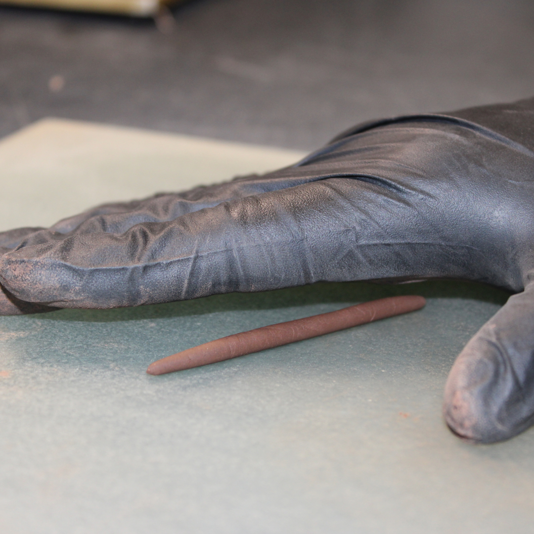 A black-gloved hand shaping a thin, brown clay rod on a light-colored surface.